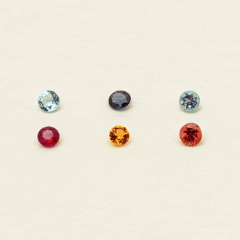 Six round-cut gemstones in assorted colors arranged in two rows on a light beige background. The top row features light blue, dark purple, and light blue stones, while the bottom row shows deep red, orange, and red-orange stones.