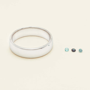 A polished silver band ring displayed on a warm cream background, with three small loose gemstones of varying colors placed to the right.