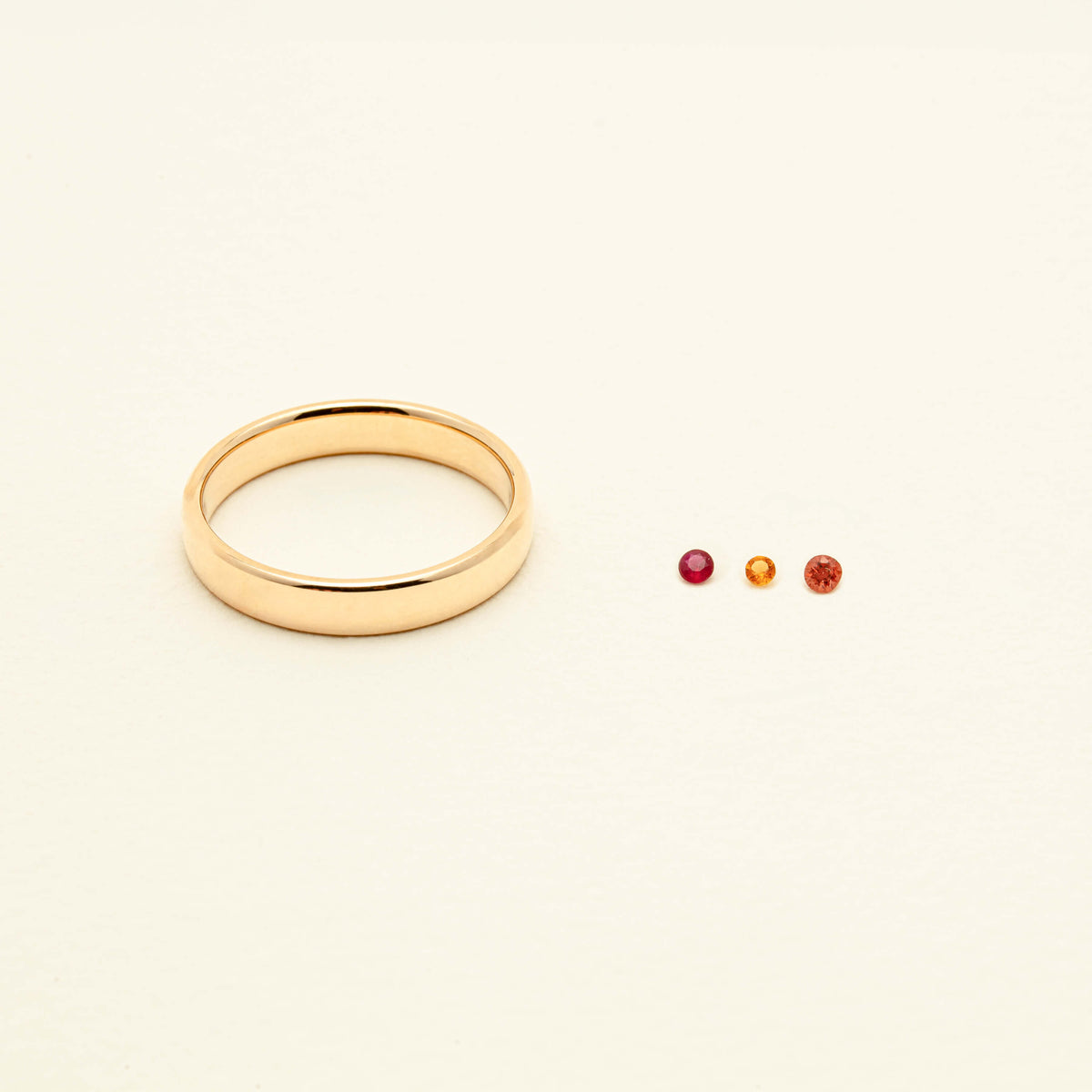 Close up of yellow gold band with three loose gemstones sitting to the right of the band. The gemstones are ruby, citrine and orange sapphire.