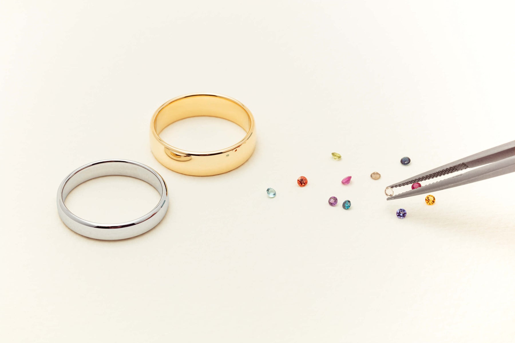 Sterling Silver and Yellow Gold plain bands with loose coloured gemstones on a neutral background. Pair of tweezers holding a morganite gemstone at the side of the image